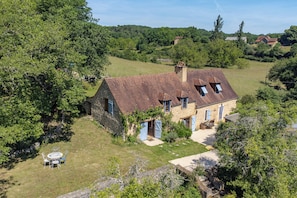 A stone house with a sloped roof, a patio with outdoor furniture, and a surrounding lush green landscape.