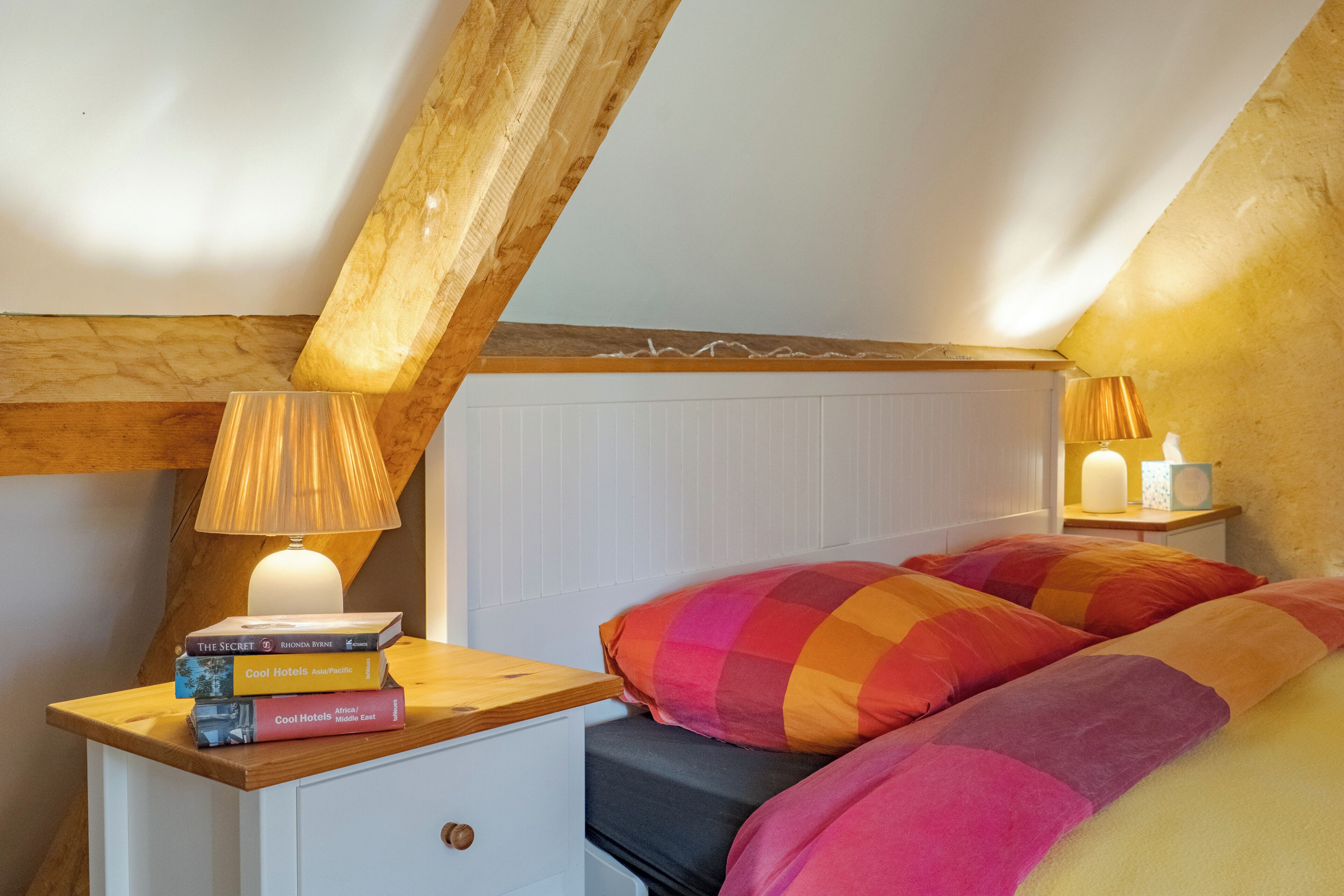 A bedroom with a wooden headboard, a bedside table with books, a lamp, and a bed with colorful bedding.