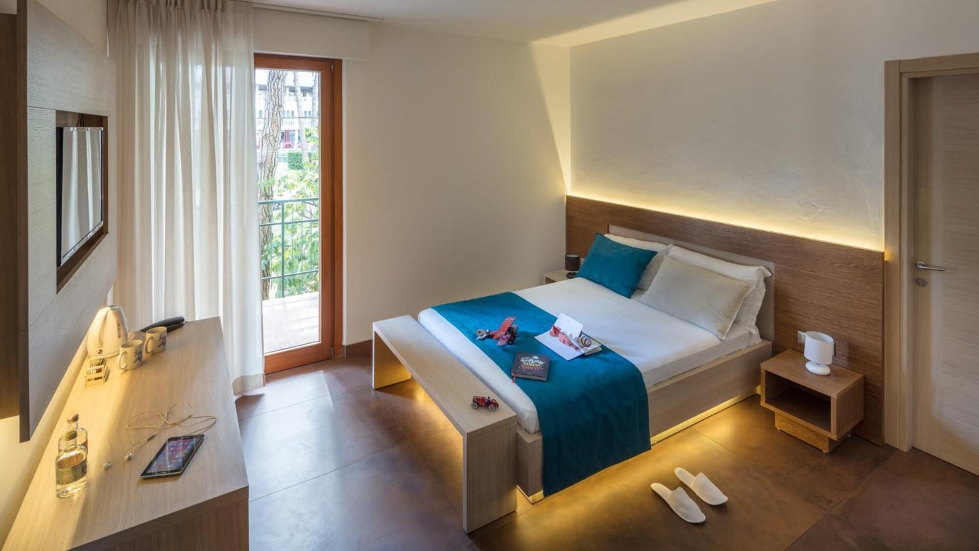 A modern hotel room with a bed, bedside tables, a desk with a chair, a window with curtains, and a view of greenery outside.