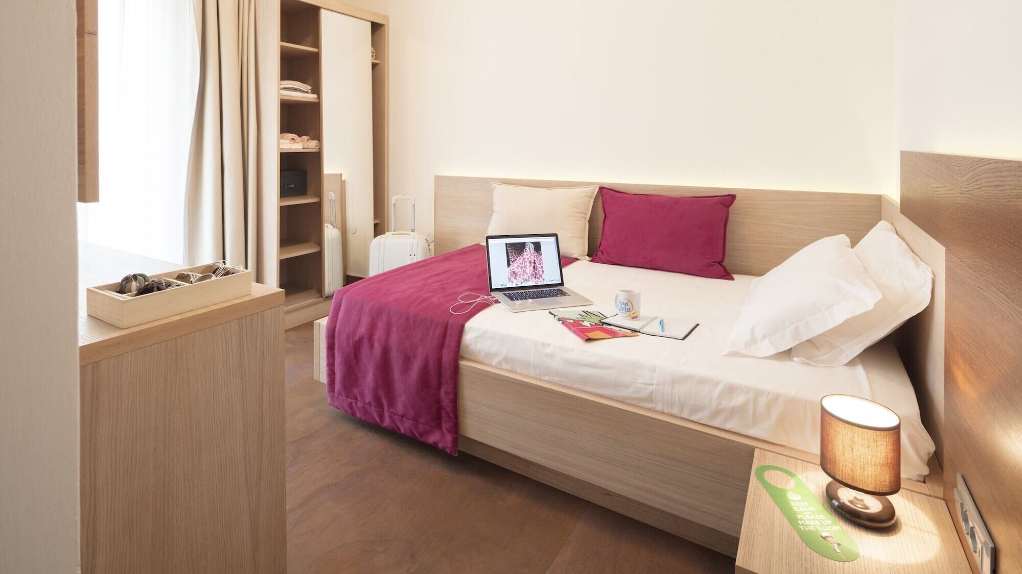 A neatly arranged hotel room with a bed, a laptop, a bedside table with a lamp, and a wooden side table with a tray.
