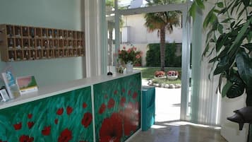 Un espace d’accueil avec un présentoir en bois où sont exposés des porte-clés, un mur peint en vert orné de fleurs rouges, une plante en pot et une vue sur un jardin avec un palmier.