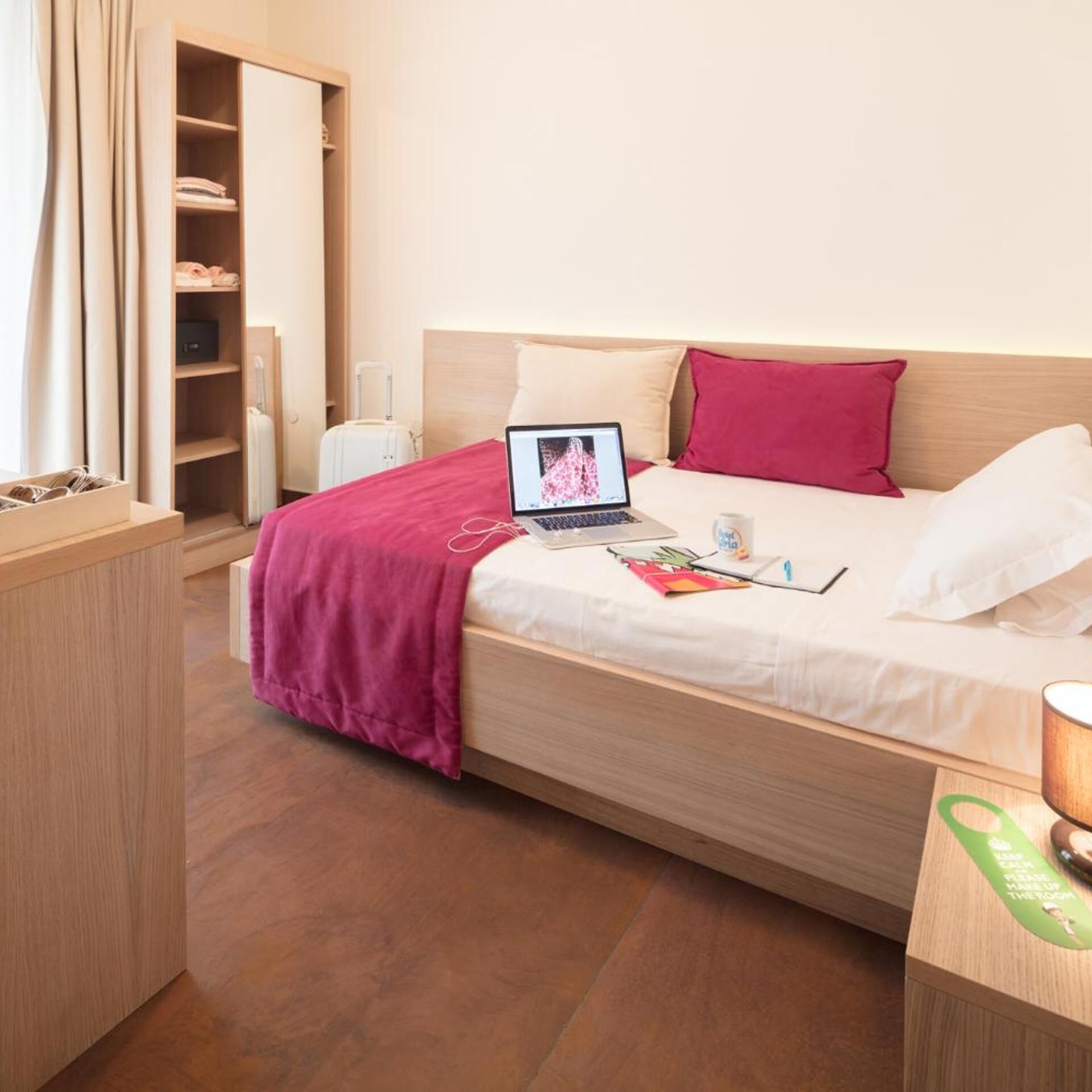 A neatly made bed with a laptop, a red pillow, and a white pillow. There is a wooden wardrobe with a mirror and a small table with a green sign.