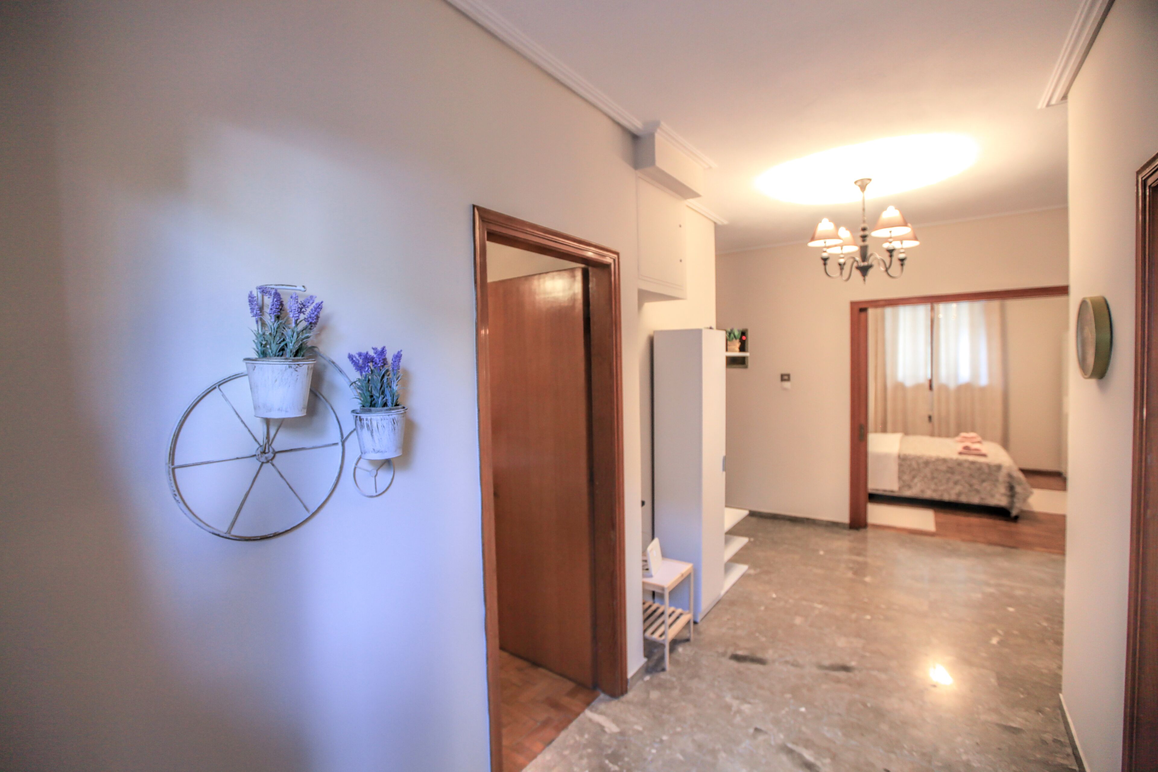 A hallway with a wooden door, a white wall, and a bed in the background.