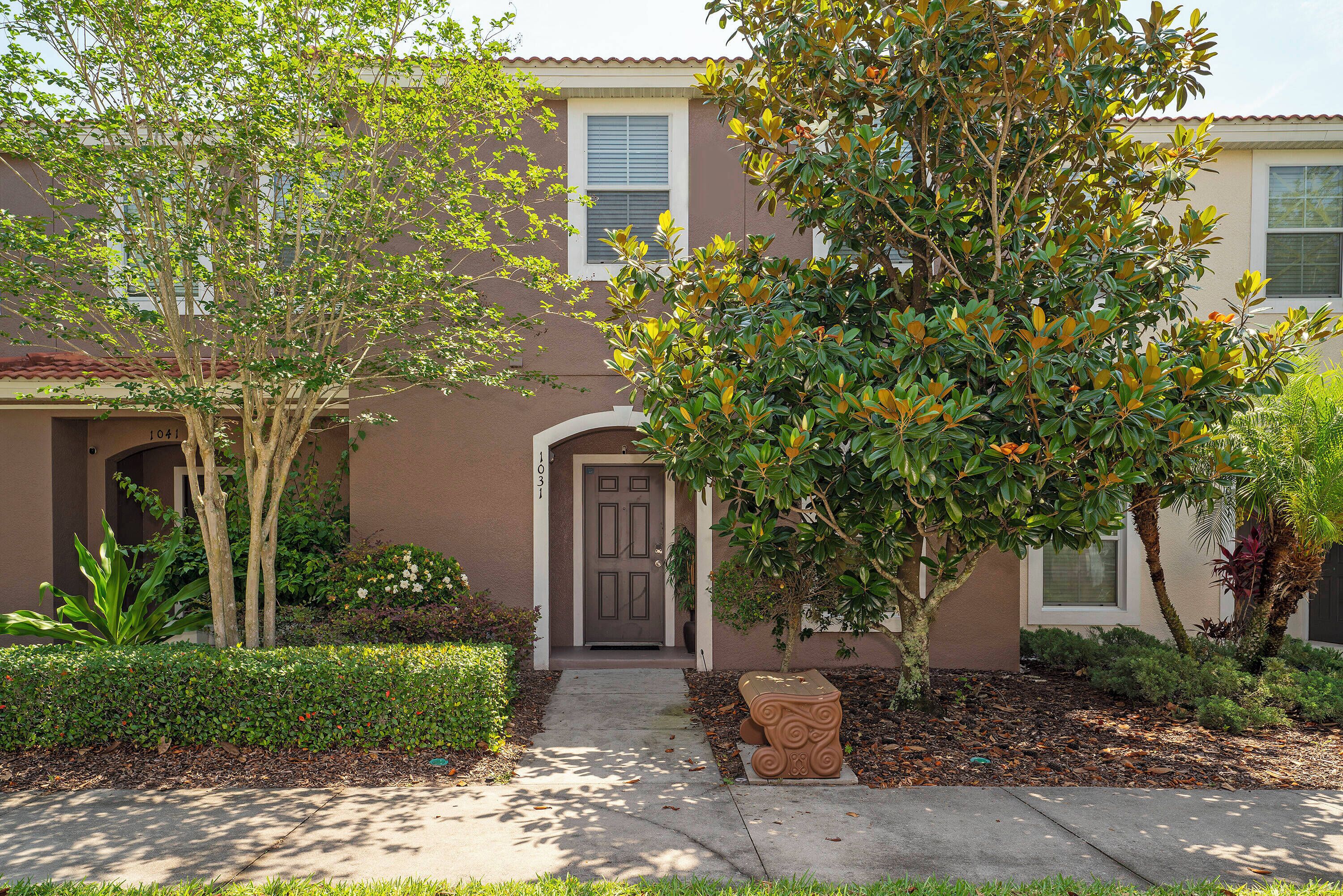 A residential building with a brown facade, a front door, and a small garden with various plants and a decorative stone planter.