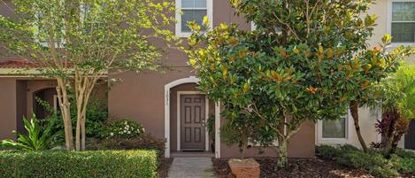 A residential building with a brown facade, a front door, and a small garden with various plants and a decorative stone planter.