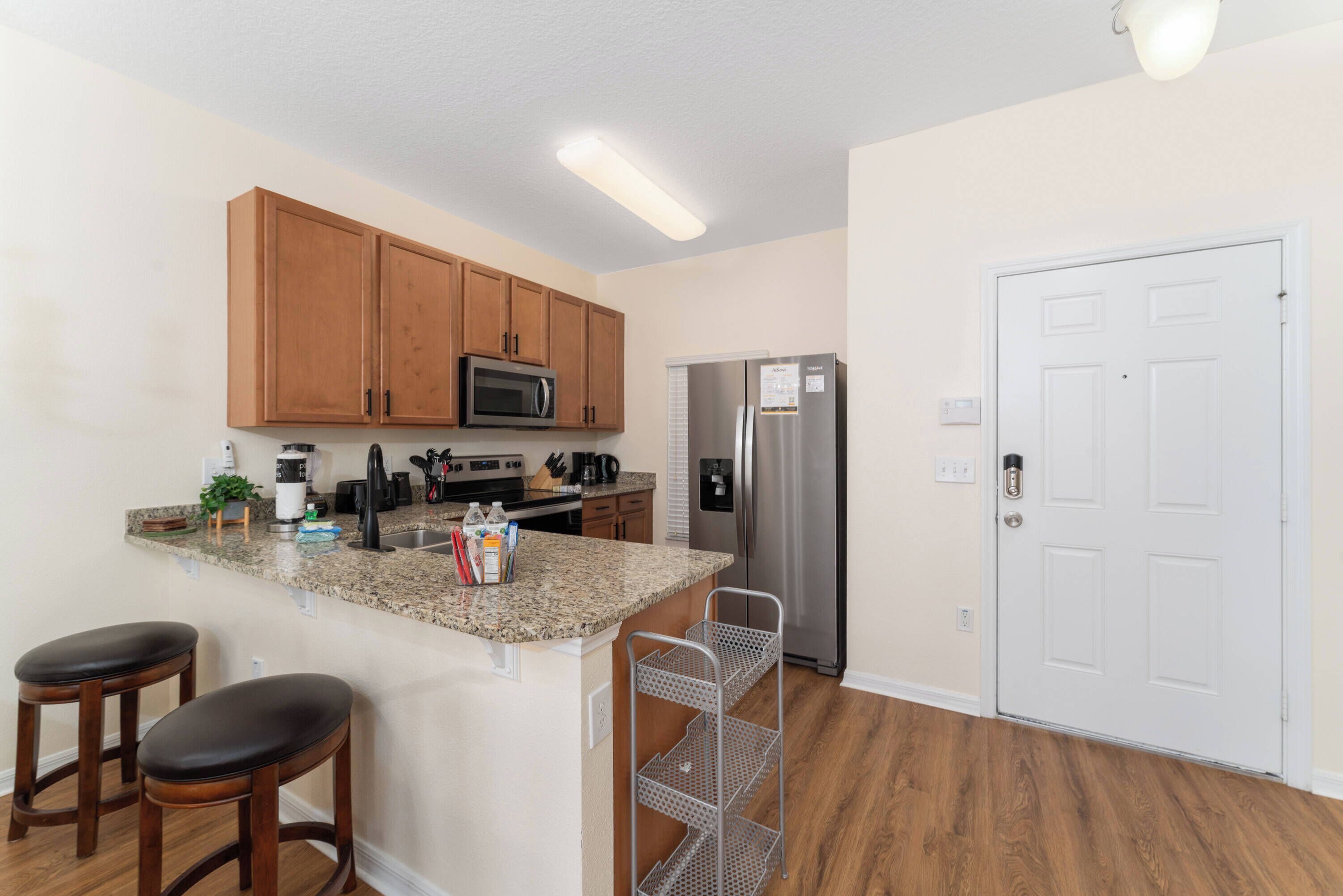 A kitchen with wooden cabinets, a stainless steel refrigerator, a granite countertop, and two bar stools.