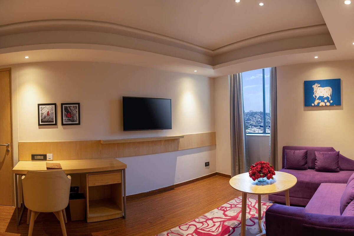 A modern hotel room with a desk, purple sofa, and a bed with a view.