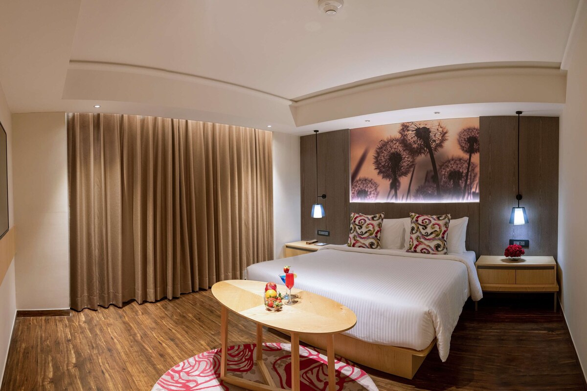 A hotel room with a bed, a bedside table with a lamp, a small round table with fruit and a drink, and a large window with curtains.