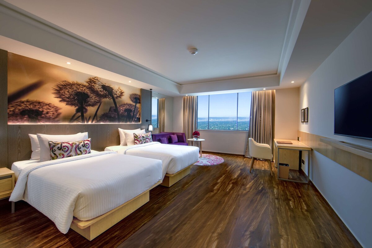 A hotel room with two beds, a desk, a chair, a TV, and a large window with a city view.