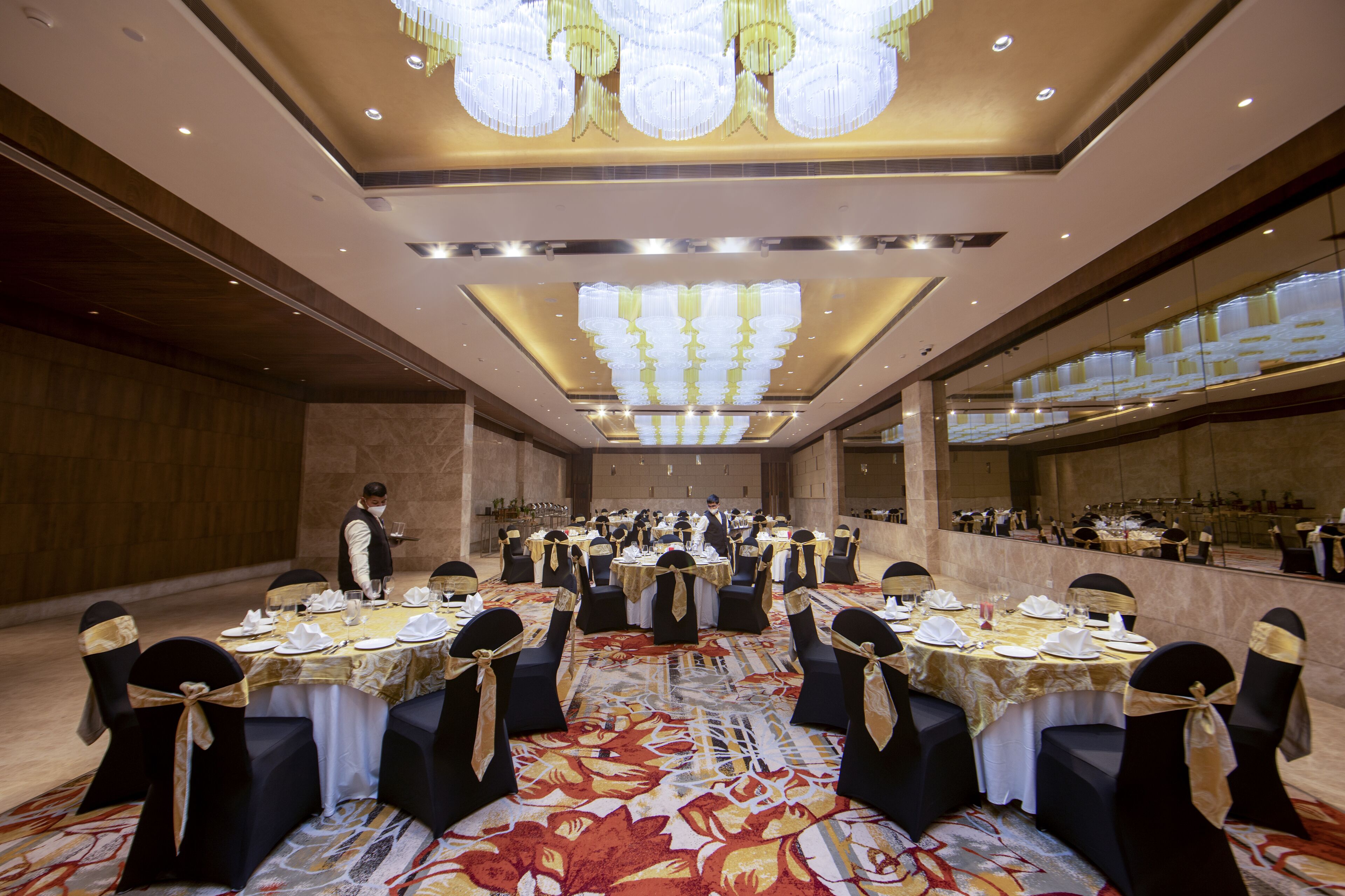 A banquet hall with round tables set for dining, a waiter in uniform, and a large chandelier.