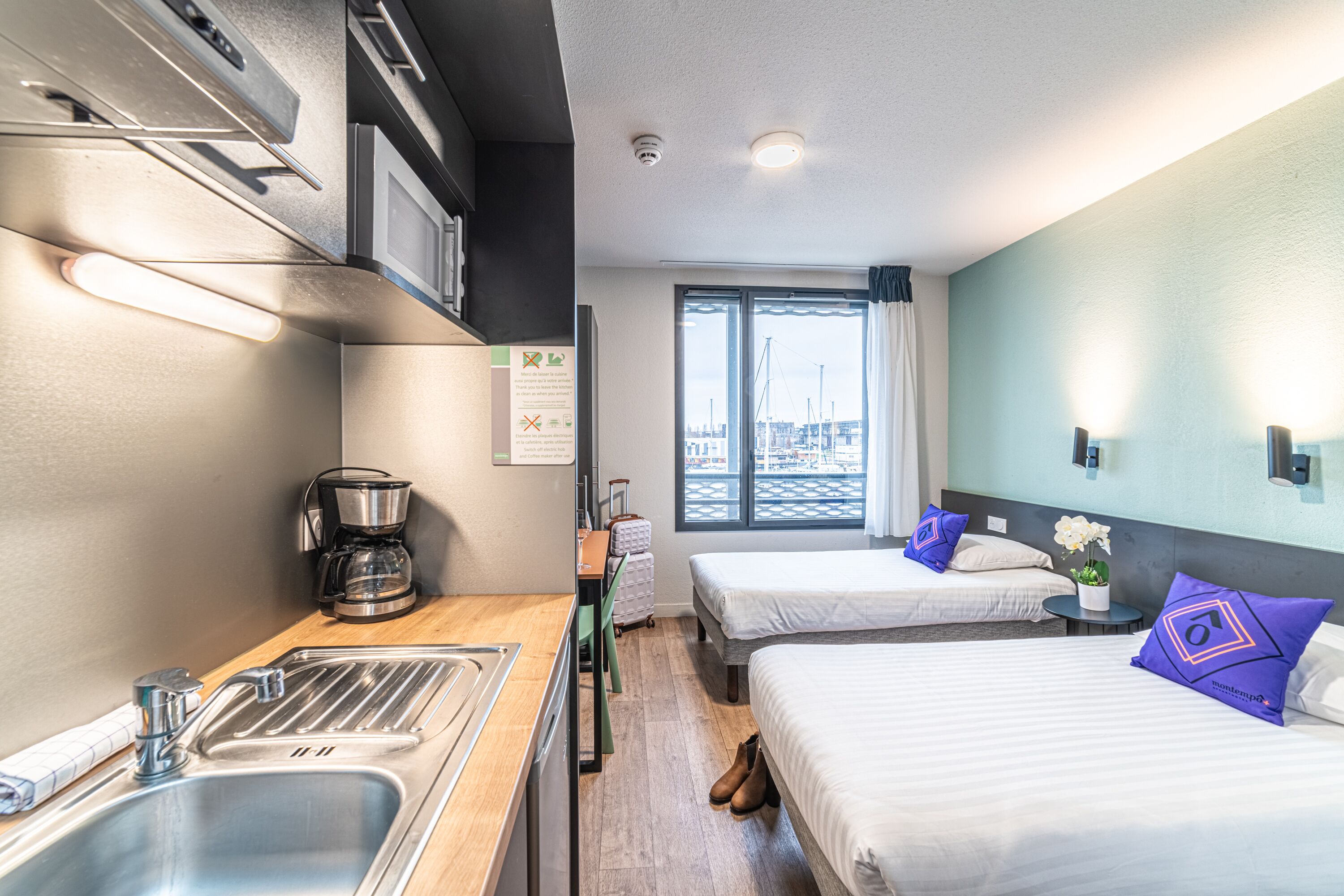Une chambre d’hôtel avec une kitchenette, deux lits, une vue sur la ville et une petite table avec une machine à café.