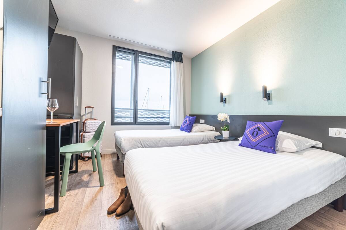 Een hotelkamer met twee bedden, een bureau, een stoel en een groot raam met uitzicht op zee.