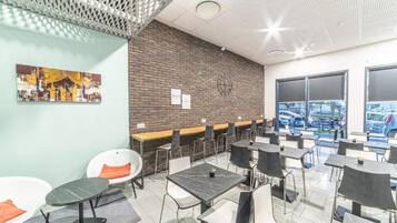 A modern dining area with tables and chairs, a brick wall with a clock, and a city view through large windows.