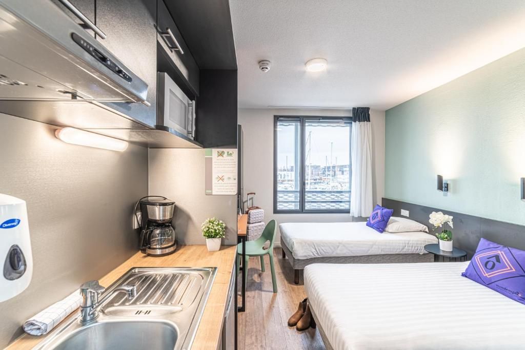 Une chambre d’hôtel compacte comprenant un coin cuisine, un lit avec un oreiller, une chaise, une petite table et une fenêtre donnant sur un paysage agréable.