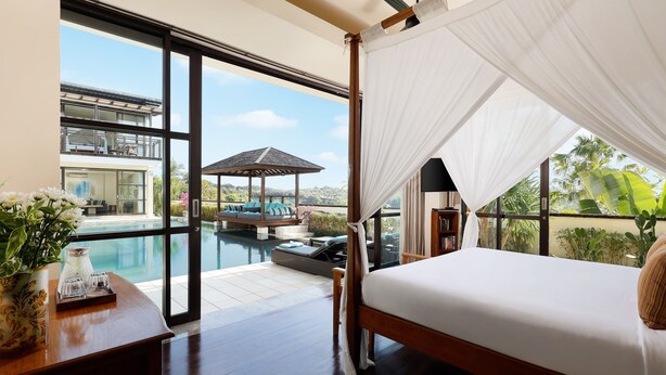A bedroom with a large window, a bed, a nightstand, and a view of a pool and outdoor seating area. - Villa Jamalu by Nakula (Jimbaran)