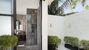A modern bathroom with a shower, a sink, and a mirror. There are potted plants and a white tiled wall. - Villa Jamalu by Nakula (Jimbaran)