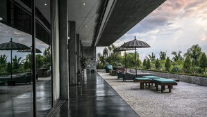 A modern outdoor lounge area with seating and umbrellas, a pathway leading to a covered entrance, and a view of tropical vegetation. - Villa Mana by Elite Havens (Canggu)