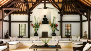 A traditional room with a wooden ceiling, a central Buddha statue, white sofas, and potted plants.