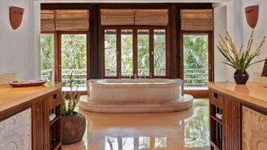 A bathroom with a large bathtub, wooden cabinets, and a view of lush greenery through large windows.