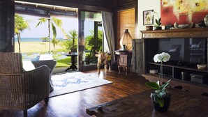 A room with a large window overlooking a beach, a fireplace, and a dining area with a view.