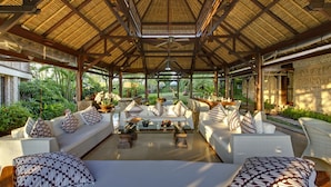 A spacious, open-plan lounge area with high ceilings and a thatched roof, featuring comfortable seating and a central coffee table.