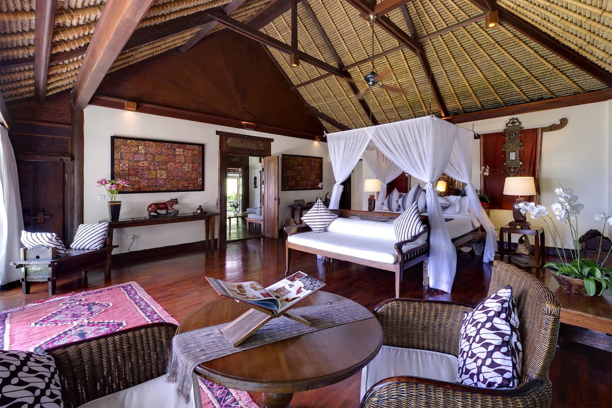 A spacious room with a high ceiling, a bed with a canopy, wicker furniture, and a large rug.