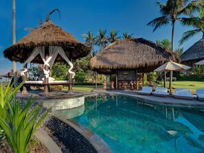 A pool area with thatched-roof structures, lounge chairs, and a clear blue pool.