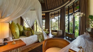 A room with a bed under a canopy, a wooden table, a wicker chair, and large windows with a view of greenery. - Taman Ahimsa by Elite Havens (Cemagi)