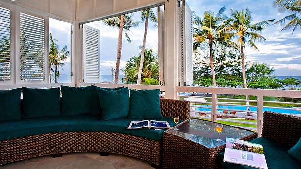A covered outdoor seating area with a wicker sofa and a glass table, overlooking a pool and palm trees. - Villa Puri Nirwana (Gianyar)