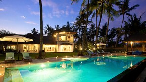 A swimming pool with lounge chairs and umbrellas, surrounded by palm trees and a two-story building with outdoor lighting. - Villa Puri Nirwana (Gianyar)