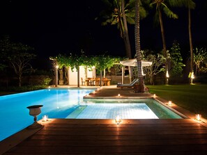 A nighttime pool area with a wooden deck, lounge chairs, and a covered seating area with candles. - Villa Puri Nirwana (Gianyar)
