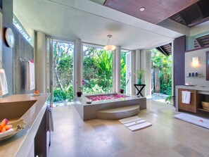 A modern bathroom with a large bathtub, a sink, and a shower area. There are plants and flowers around the room, and a view of the outdoors. - Villa Samadhana (Sukawati)