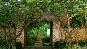 A stone Buddha statue under a thatched roof, surrounded by lush greenery. - Villa Samadhana (Sukawati)