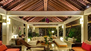 A modern living room with a wooden ceiling, a central seating area with sofas, a coffee table, and a large window overlooking a garden. - Villa Samadhana (Sukawati)