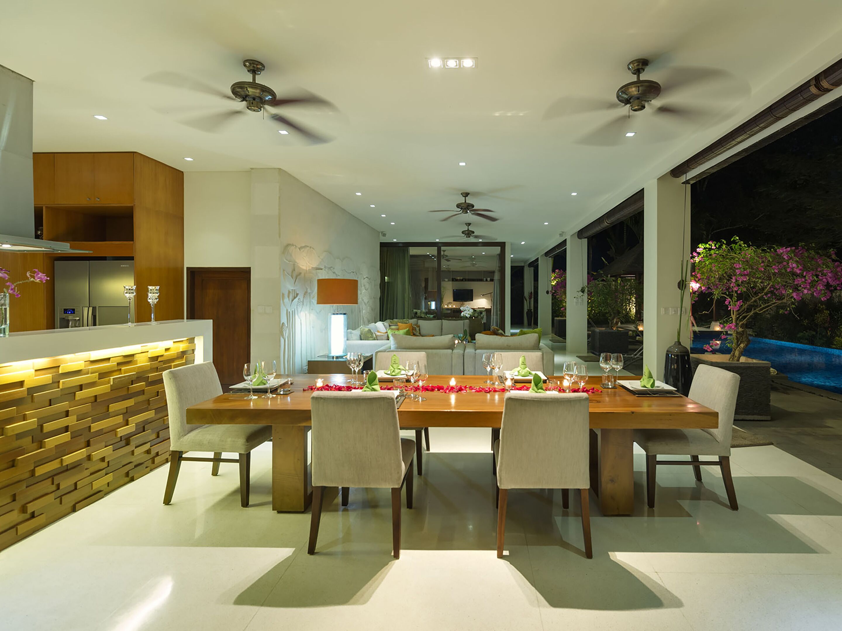 Un comedor moderno con una mesa de madera dispuesta para seis personas, rodeada de sillas. Hay ventiladores de techo, iluminación empotrada y vistas al exterior.