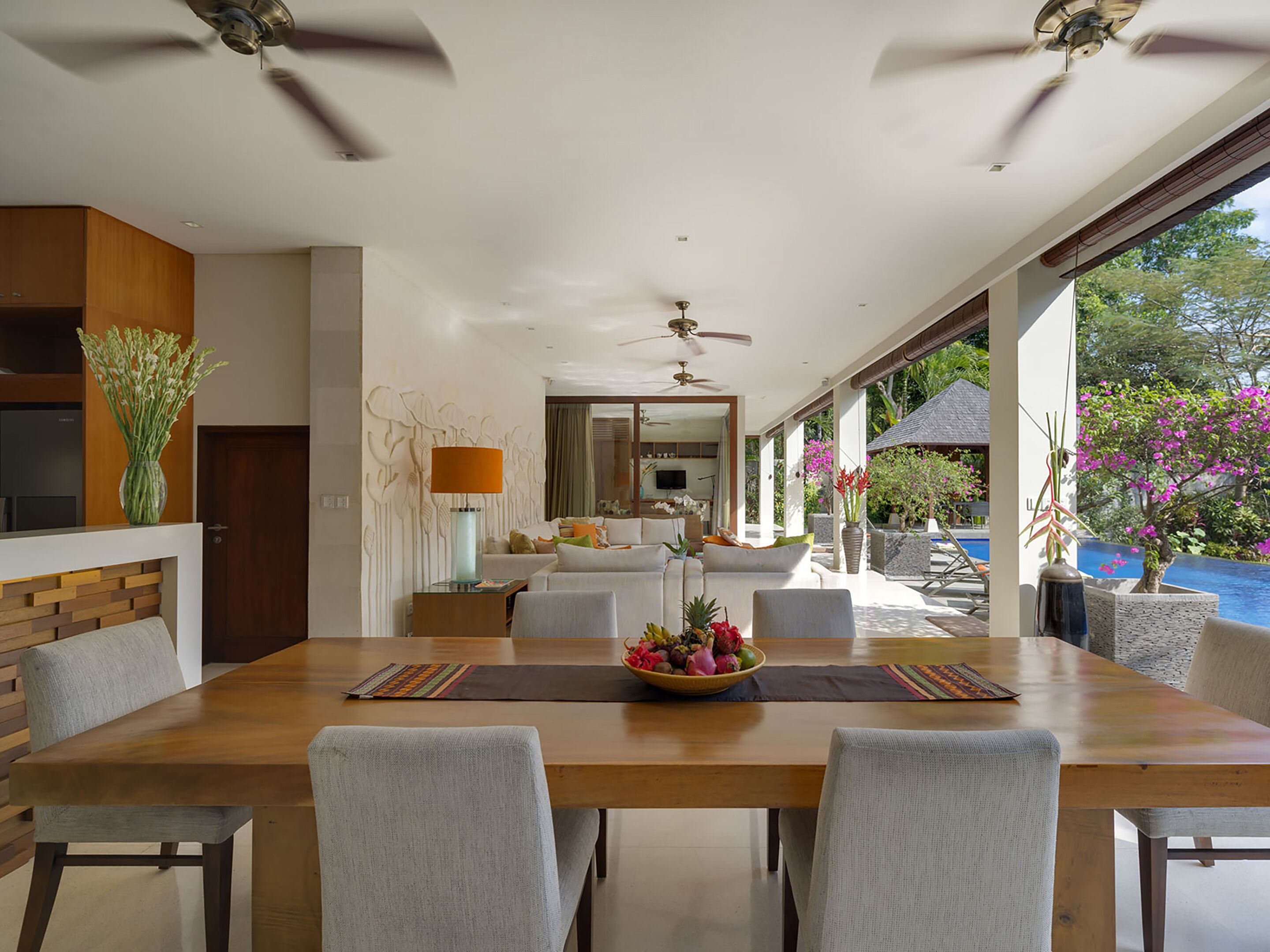 A modern dining area with a wooden table, grey upholstered chairs, a central fruit bowl, and a ceiling fan.
