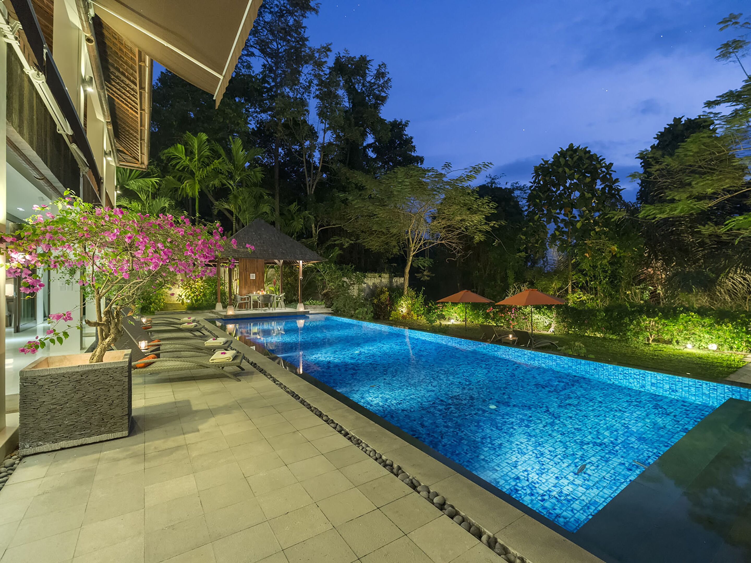 A swimming pool with a tiled deck, surrounded by lush greenery and a gazebo.