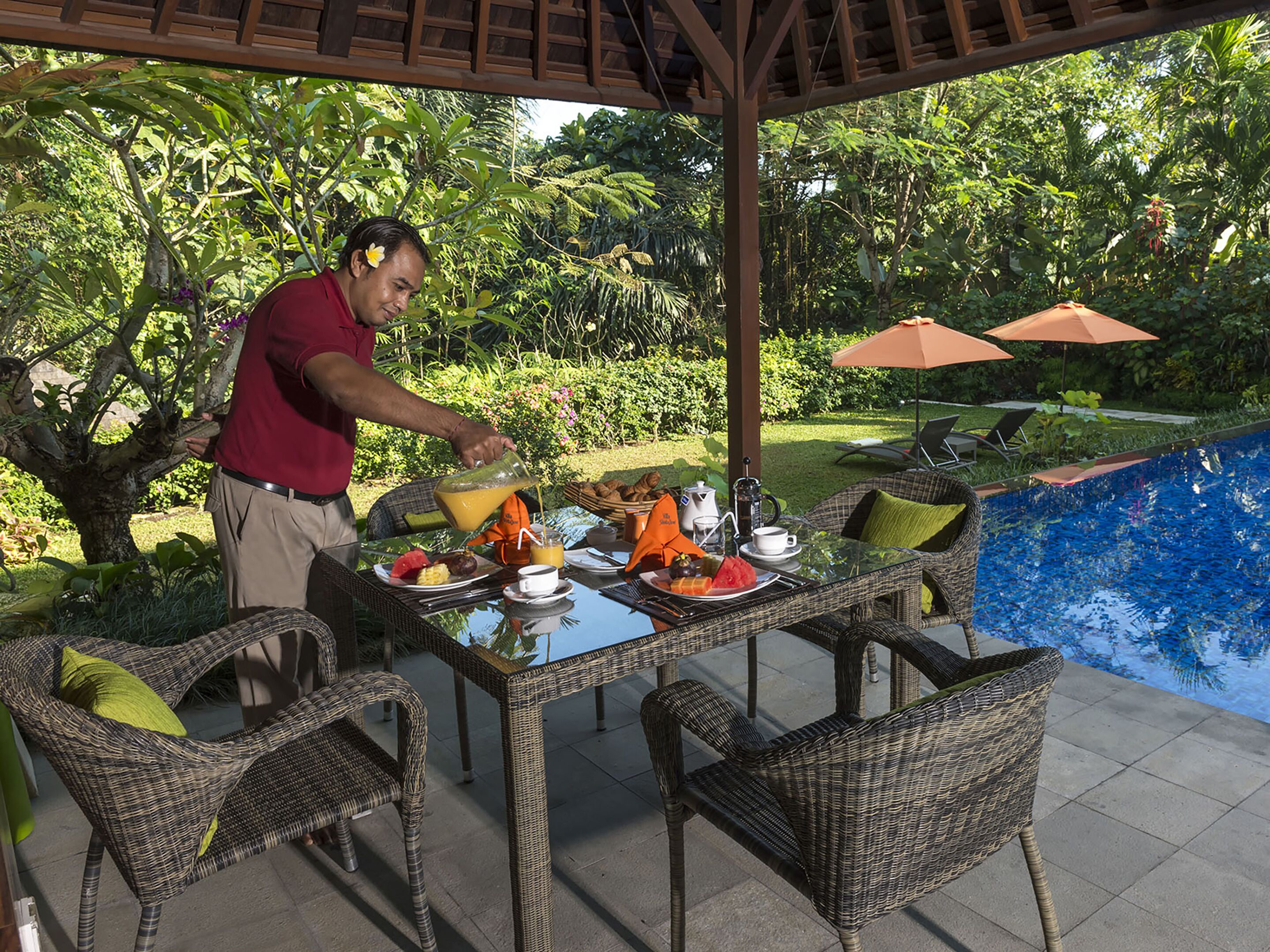 A covered outdoor dining area with wicker furniture, a pool, and lush greenery in the background.