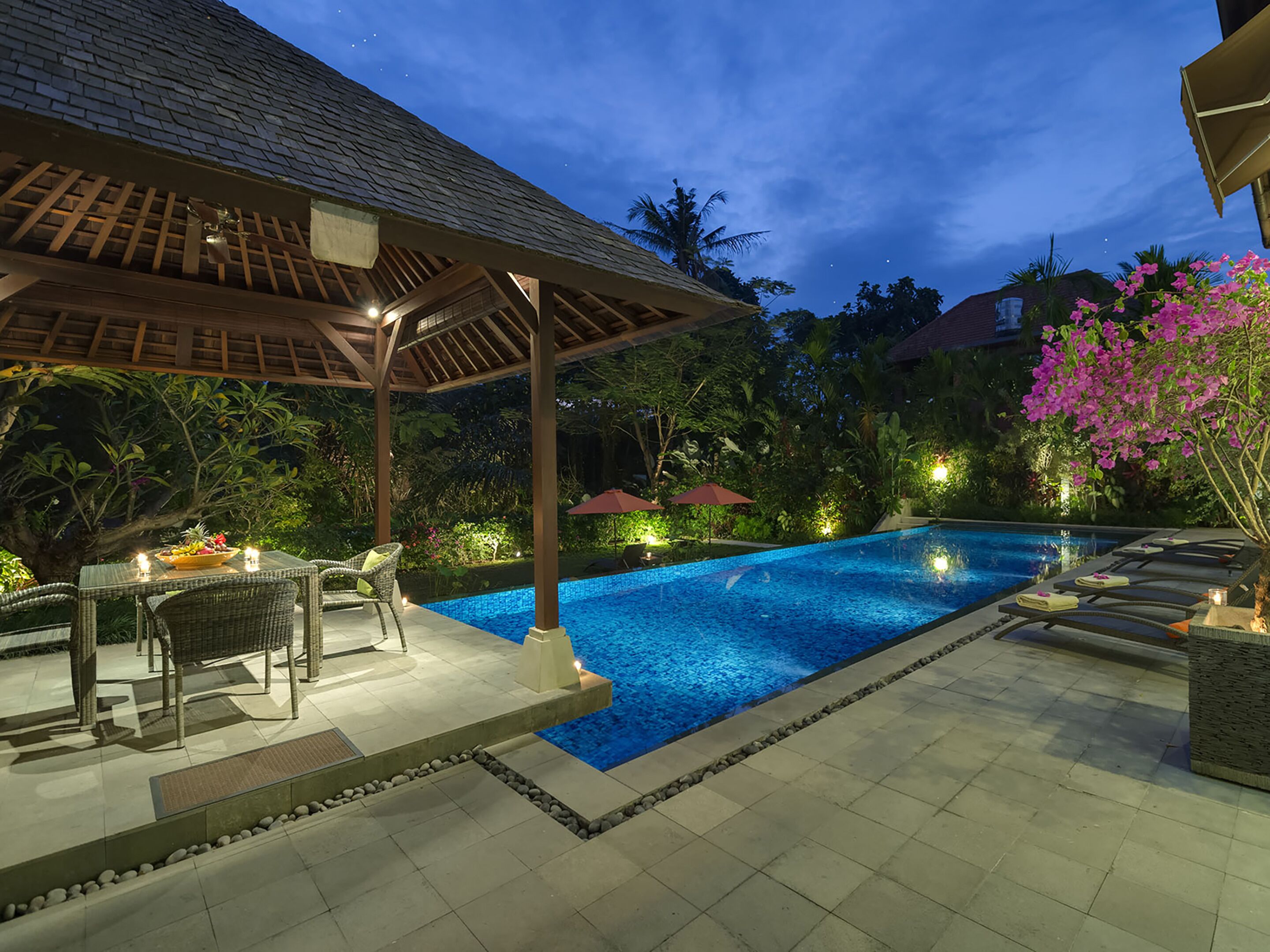 Una piscina con pérgola y sillones de descanso, rodeada de abundante vegetación y un cielo nocturno.