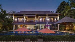Una casa de dos pisos con piscina, sillones de descanso y sombrillas.