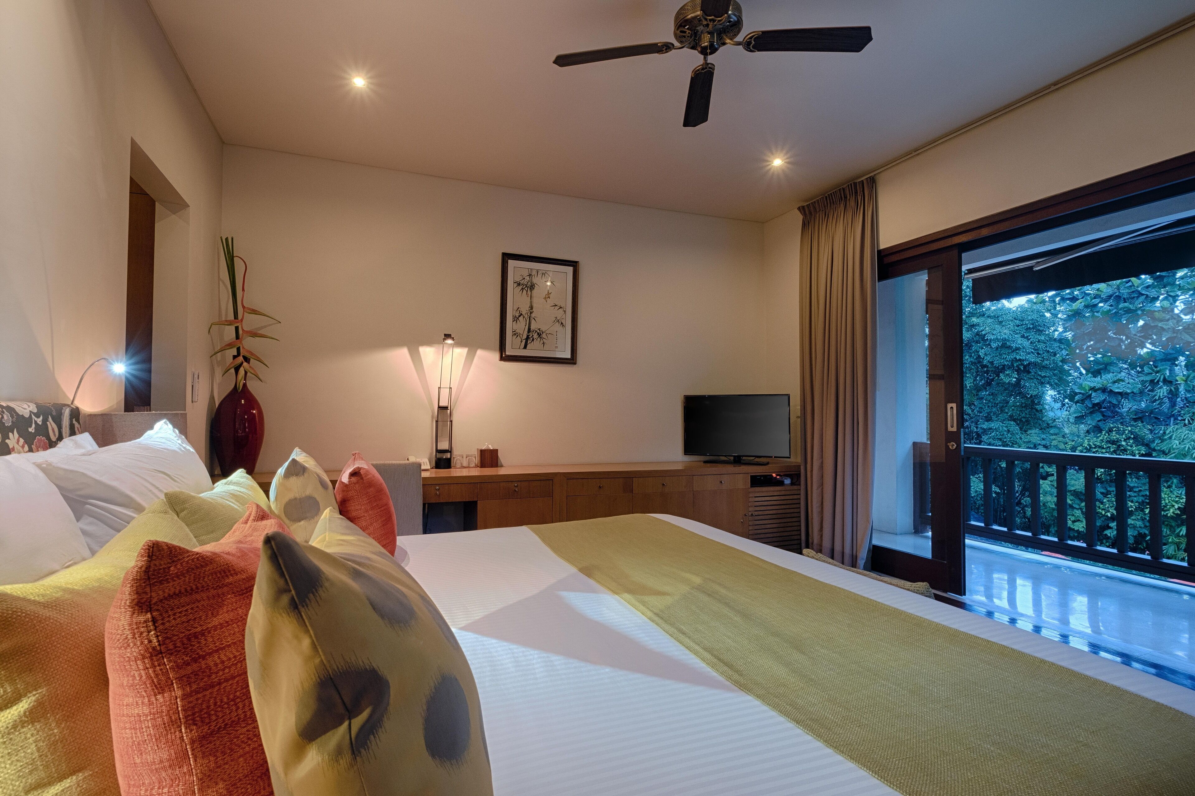 Et hotelværelse med seng, fjernsyn, skrivebord, lampe, plante og balkon med udsigt til træer.