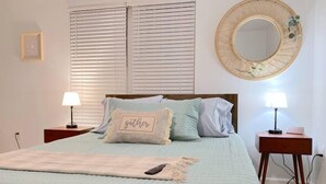 Ein Schlafzimmer mit einem Bett, zwei Nachttischen, einem runden Spiegel und einem Fenster mit Jalousien.