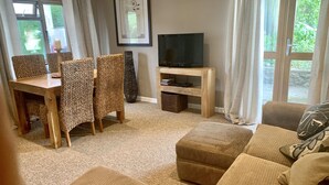 A living room with a wooden dining table, wicker chairs, a sofa, a TV on a wooden stand, and a large window with curtains. - The Stables, Lovely Country Apartment in a Converted Barn (Wells)