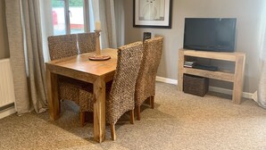 A dining area with a wooden table and woven chairs, a television on a wooden stand, and a framed artwork on the wall. - The Stables, Lovely Country Apartment in a Converted Barn (Wells)