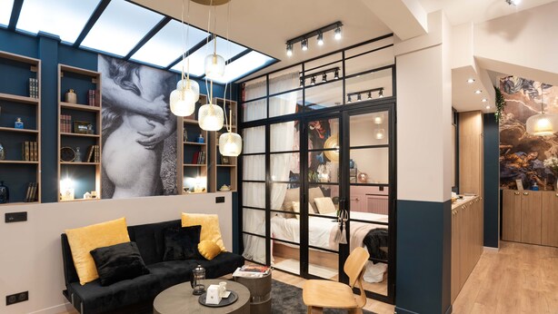 Une pièce moderne avec un canapé noir, des coussins jaunes, une table basse ronde et une chaise en bois. On y trouve des étagères remplies de divers objets et une grande fenêtre offrant une belle vue.