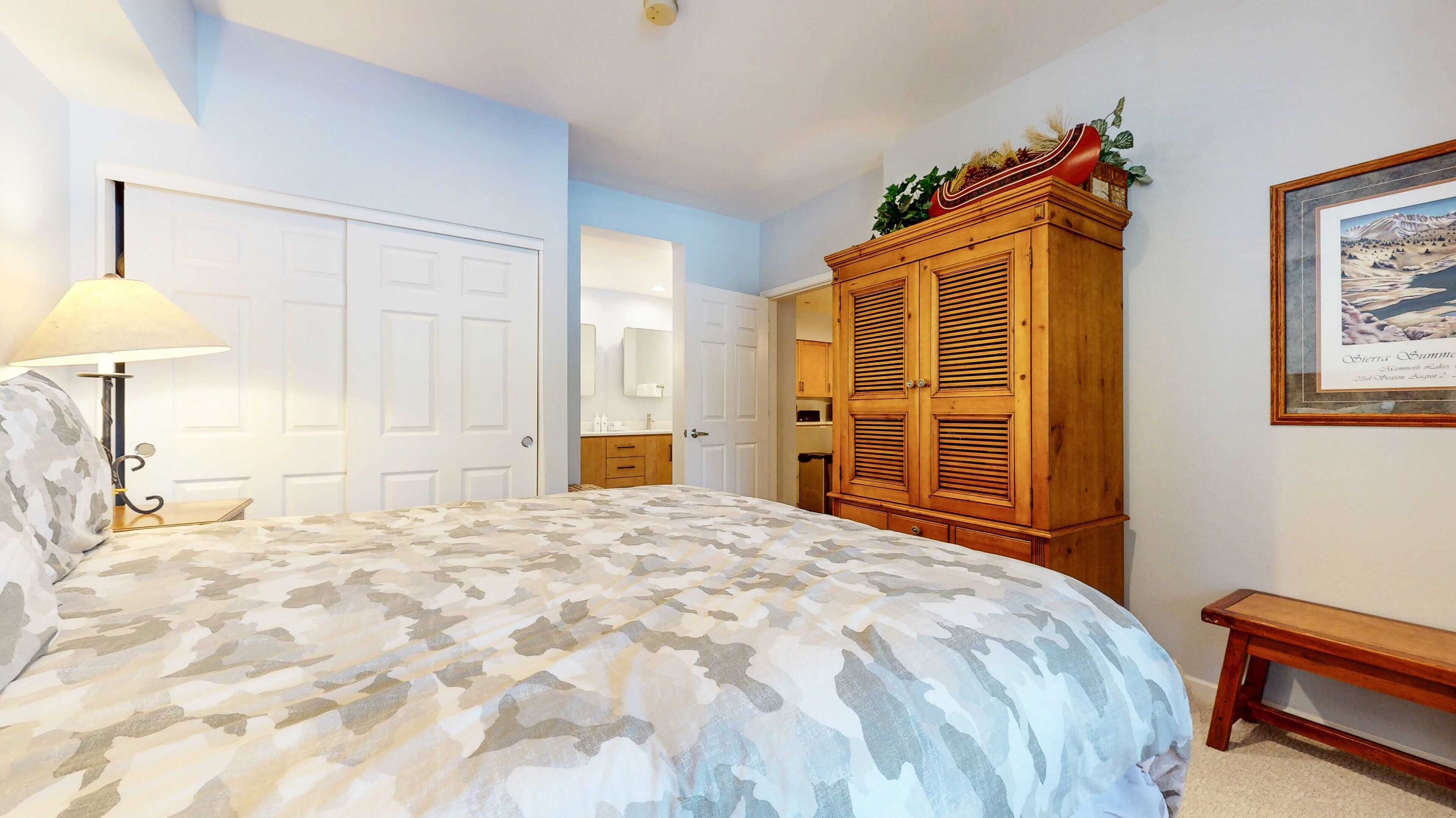 A bedroom with a bed, a wooden wardrobe, a bench, and a framed picture on the wall.