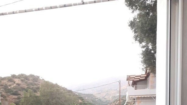 Un balcon avec une table et des chaises, donnant sur un paysage vallonné où l’on aperçoit des bâtiments au loin.