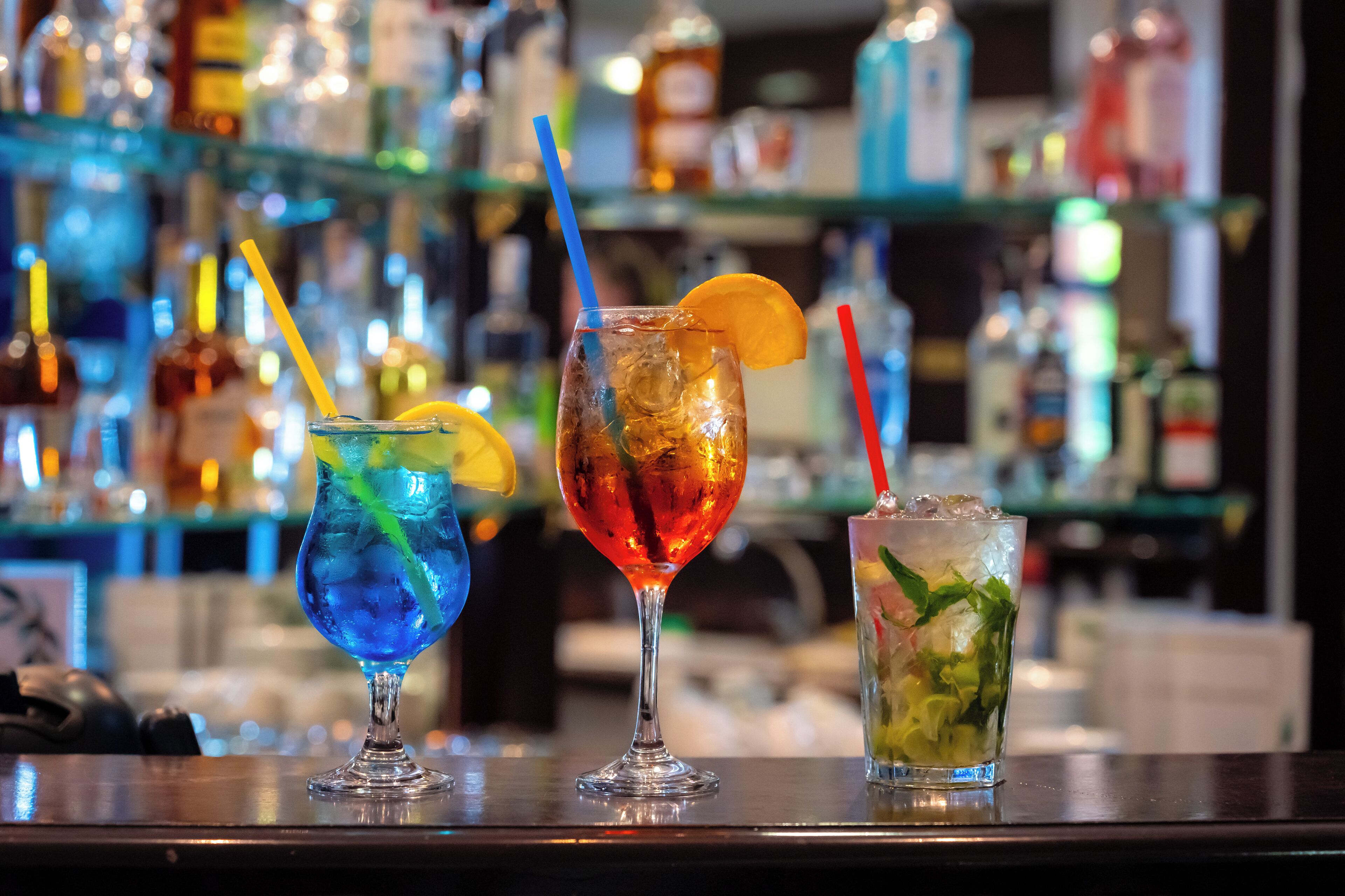 Three cocktails on a bar counter with a blurred background of shelves stocked with bottles.