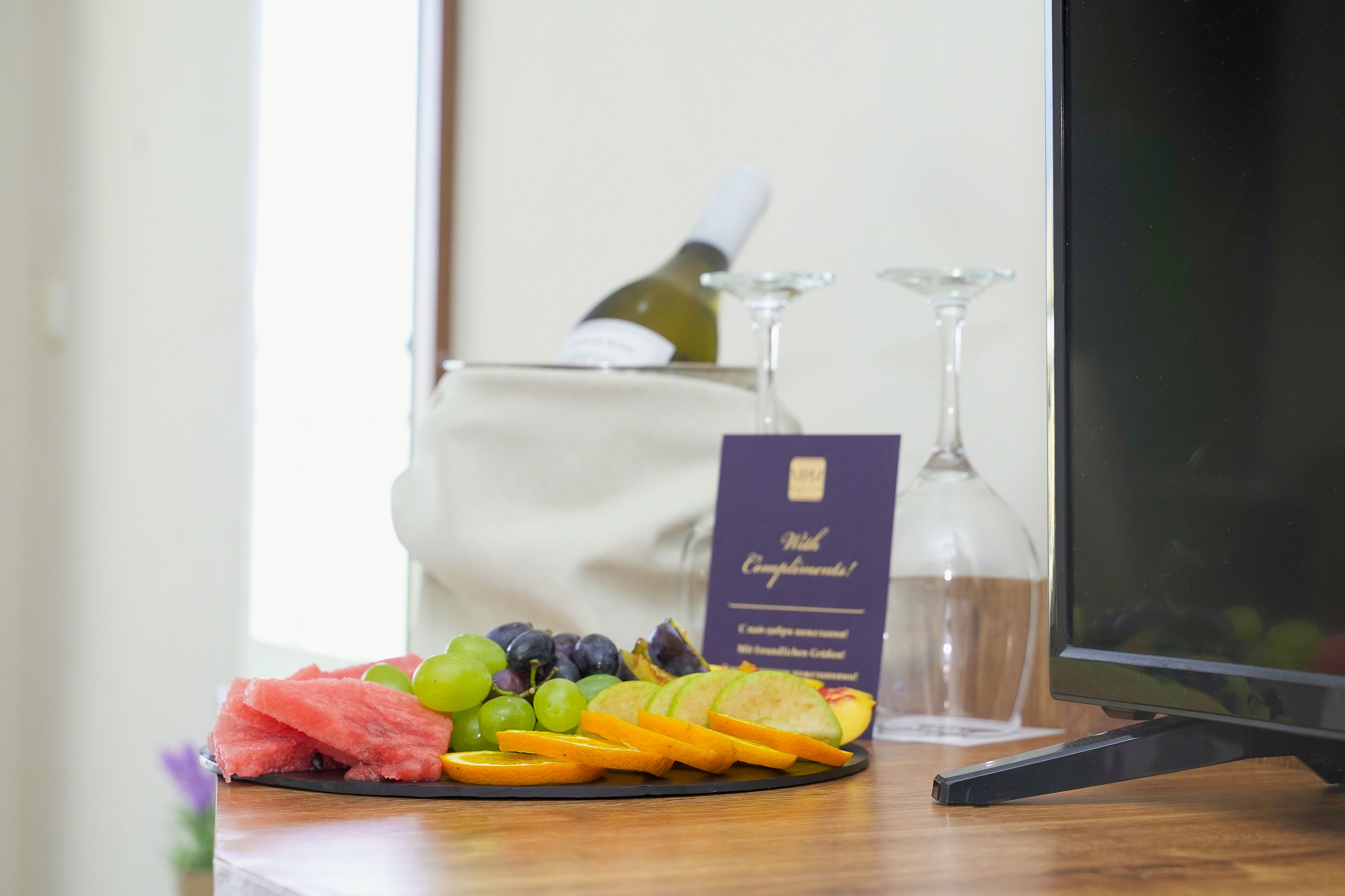 A fruit platter with watermelon, grapes, and orange slices on a black plate, a wine bottle, and two wine glasses in the background.