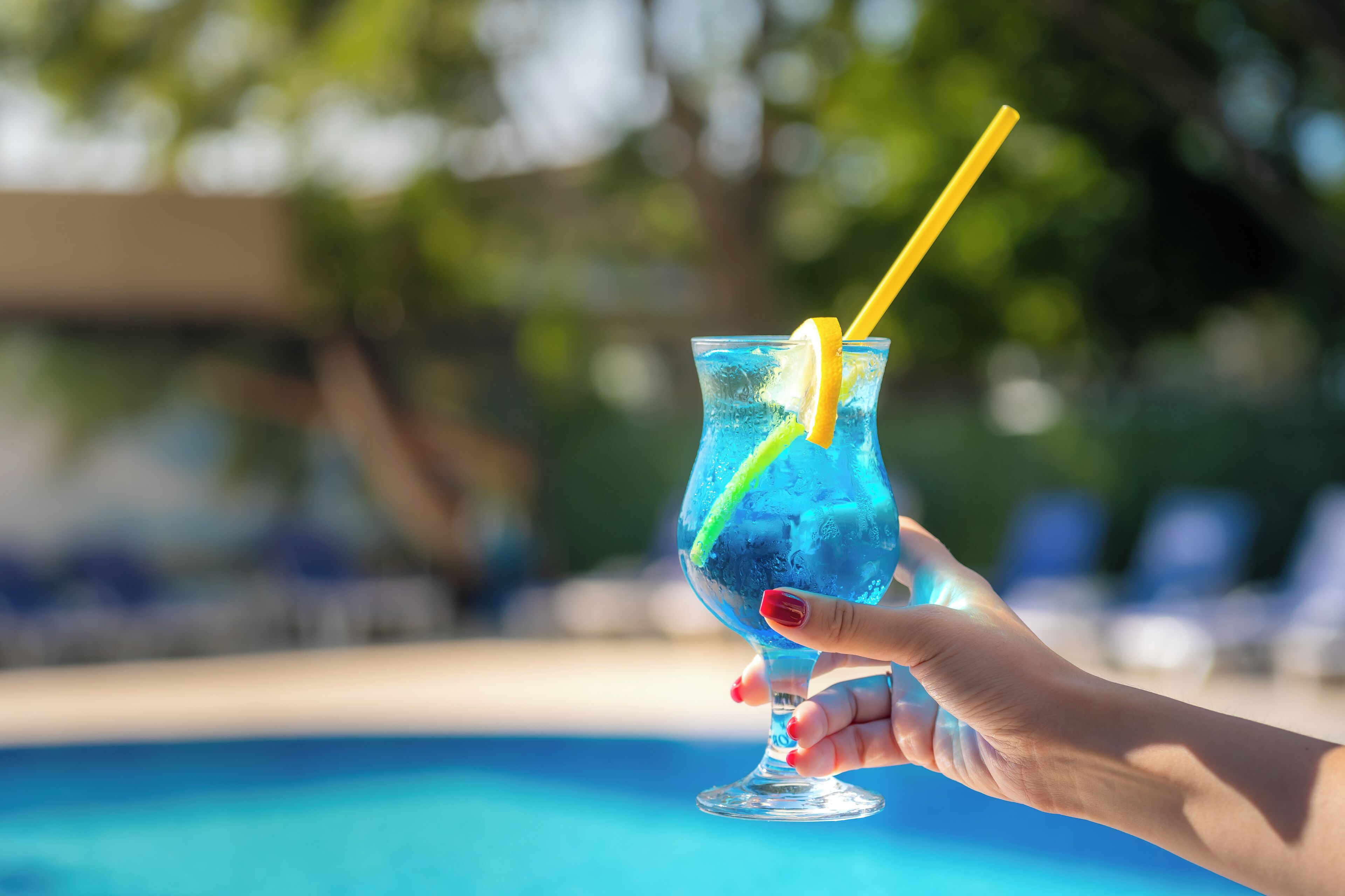 A hand holding a blue cocktail with a lemon slice and straw, near a swimming pool.